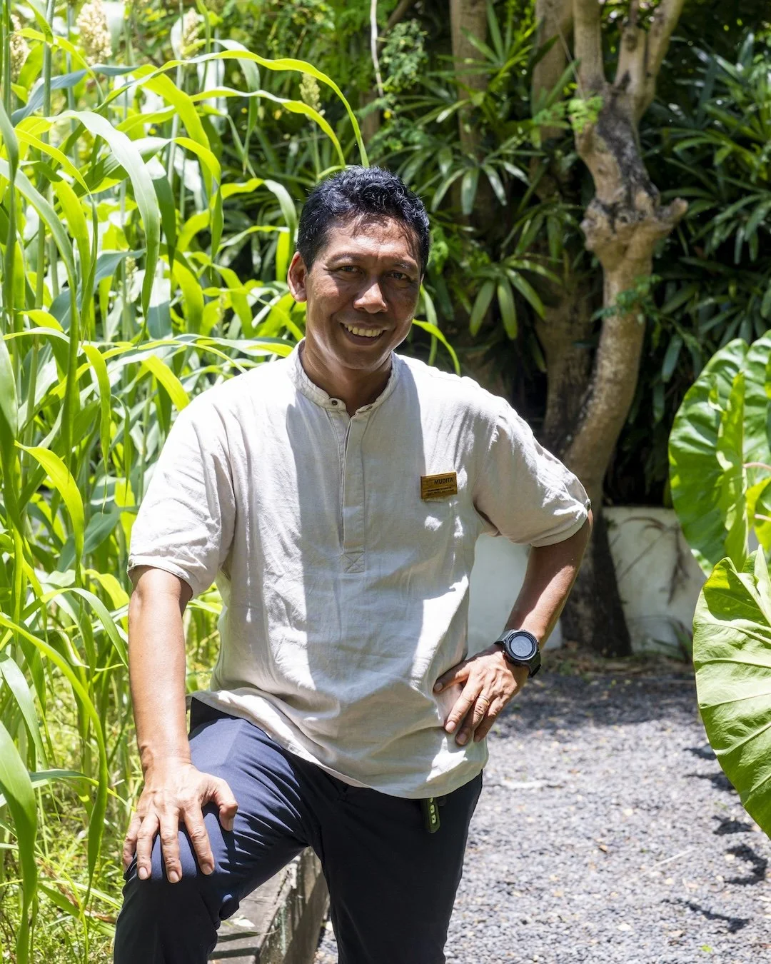A man standing outdoors in a lush garden, smiling, wearing a beige shirt with a name tag, black pants, and a black watch, with greenery and trees in the background.