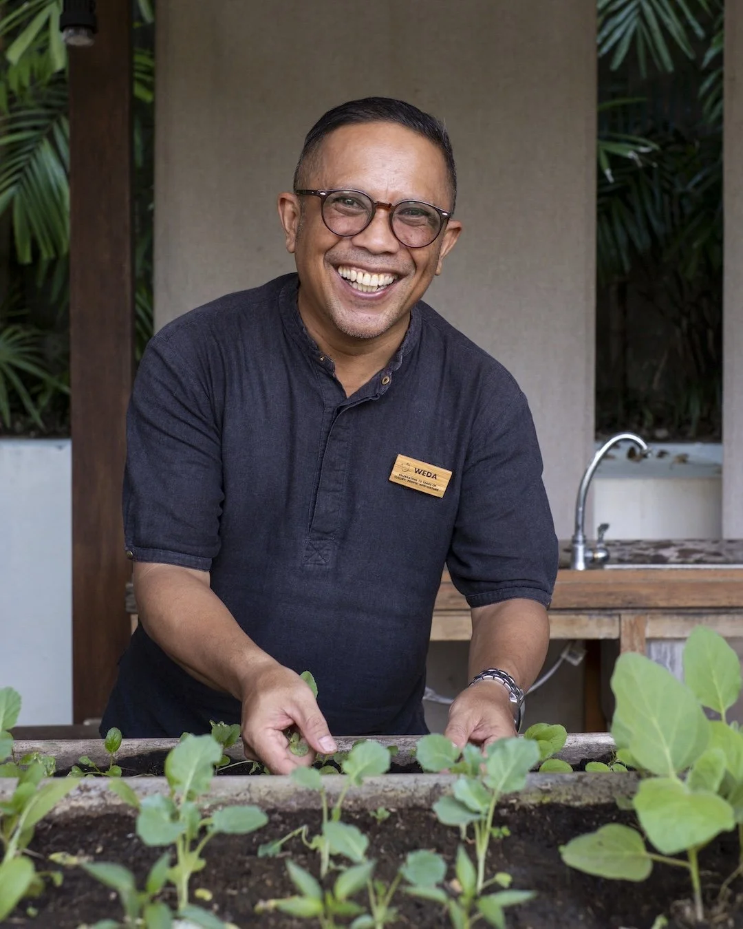A smiling man wearing glasses and a black shirt with a name tag, working in a garden with green leafy plants and soil.