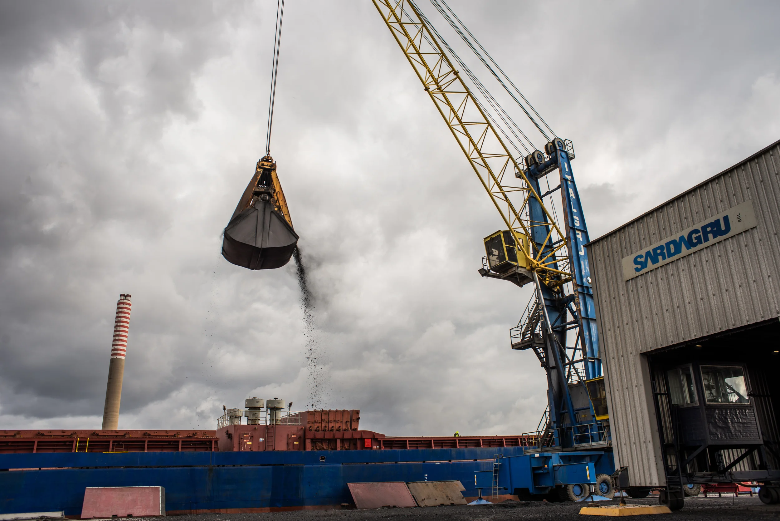 Manovre di scarico di una nave che trasporta pet-coke diretto alla Portovesme srl che lo usa come carburante per i forni Waeltz. Il suo elevato tenore di zolfo lo rende un combustibile altamente inquinante. Portovesme possiede un unico porto industriale e turistico, nel quale transitano i carichi da e per le fabbriche e i passeggeri diretti alla vicina isola di Carloforte. La minaccia della contaminazione è tale che nei giorni in cui il maestrale soffia forte la Capitaneria di Porto è costretta a interrompere lo scarico delle merci.