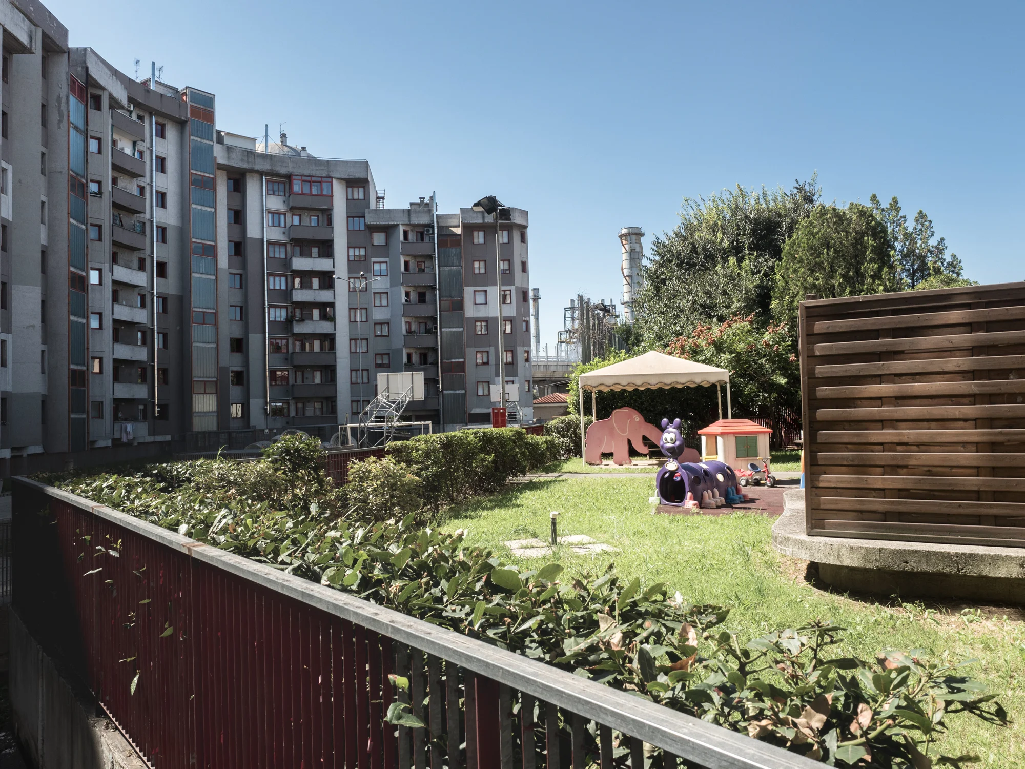 Area giochi dell'asilo nido "La Mongolfiera" nel complesso popolare di Valmaura a Trieste, 2015. Alle spalle dell'area si intravedono le "torri" della Elettra Produzione S.r.l, società privata che proprio dalla Ferriera ottiene i gas di cokeria con cui produce energia.