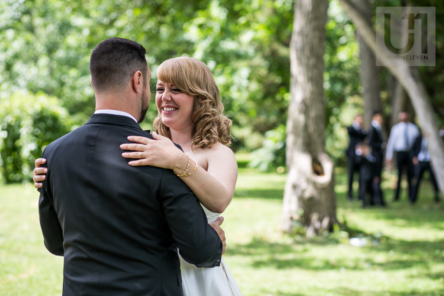 bride-smiling-embracing-groom-during-first-look-while-groomsmen-watch-in-distance