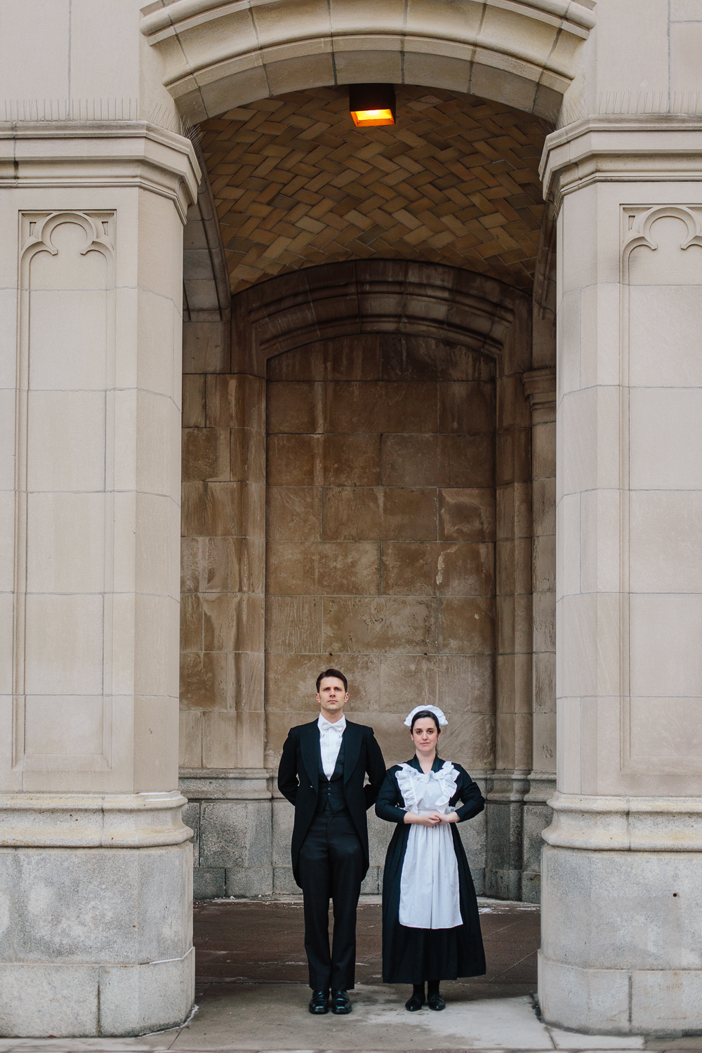 Engagement shoot of the couple posing in front of stone pillars dressed as a butler and maid