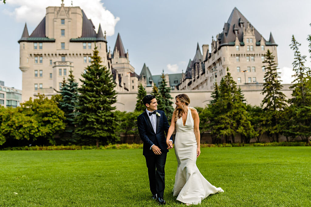 bride and groom walking on grass in front of Chateau Laurier in Ottawa while holding hands.