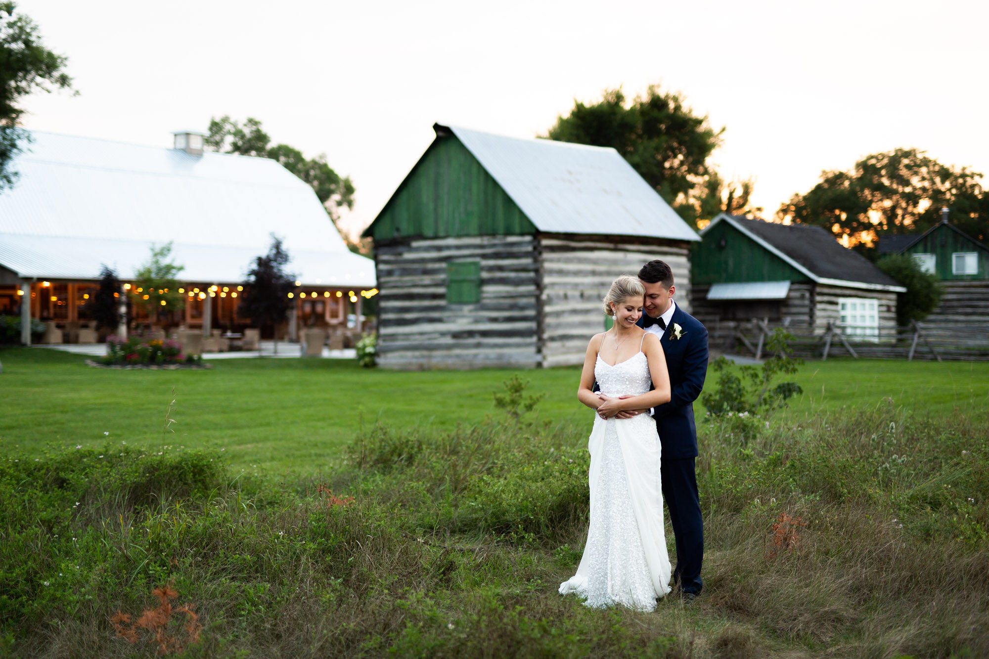 Portrait of groom holding bride from behind in front of farm buildings on Stonefields Estate property