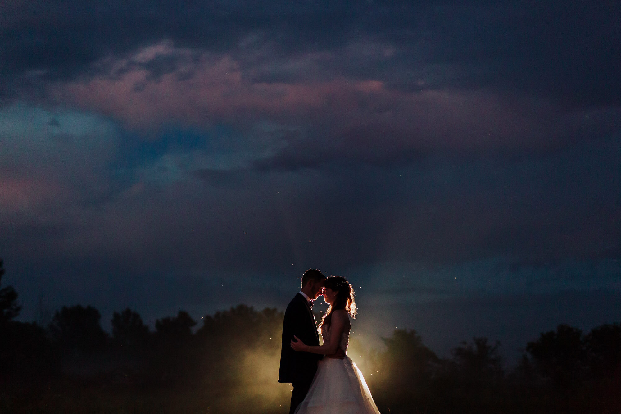 Posed portrait of bride and groom facing each other, backlit in front of a dramatic evening sky