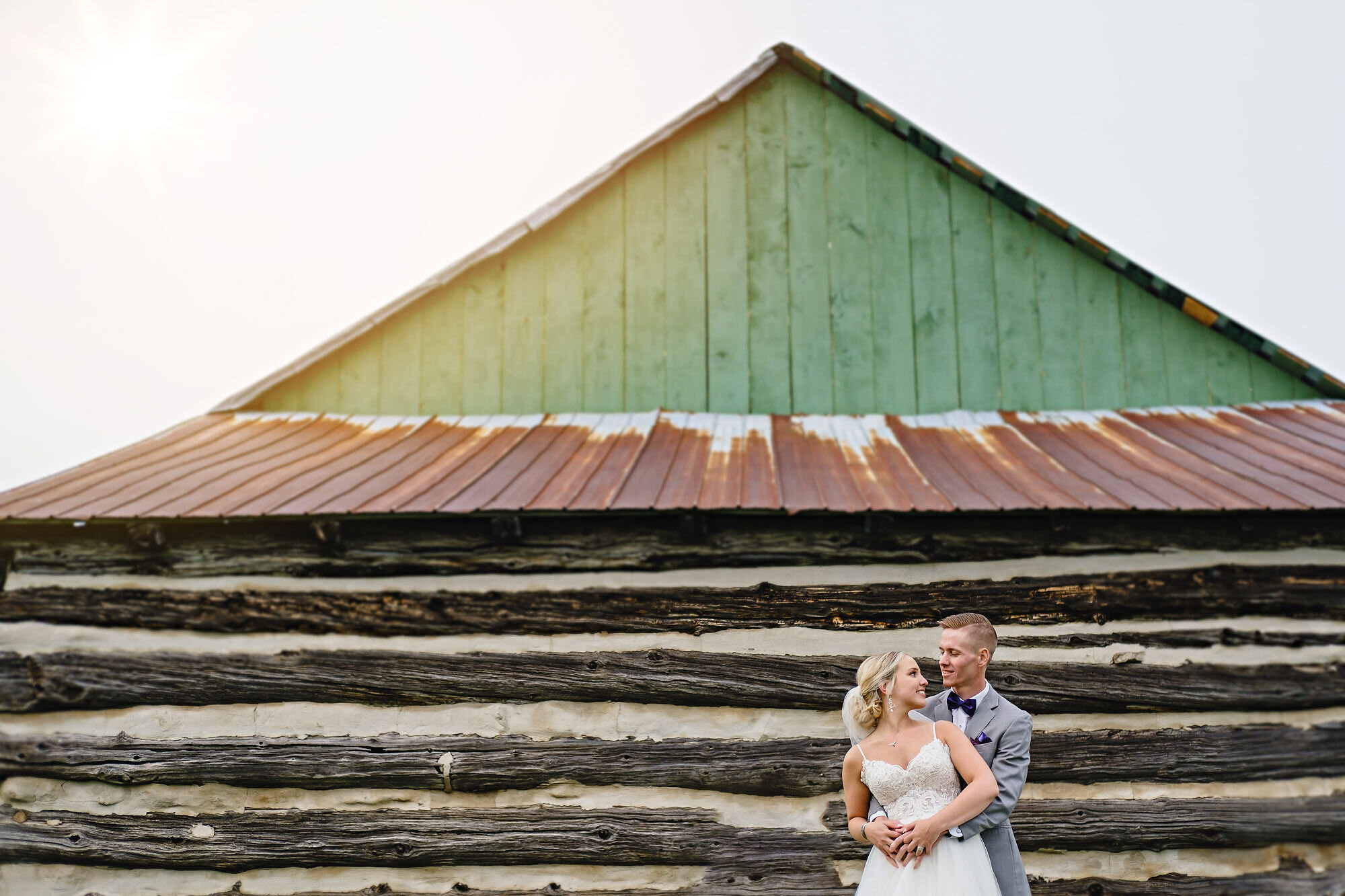 Photograph of bride and groom standing in an embrace in front of log cabin with green roof