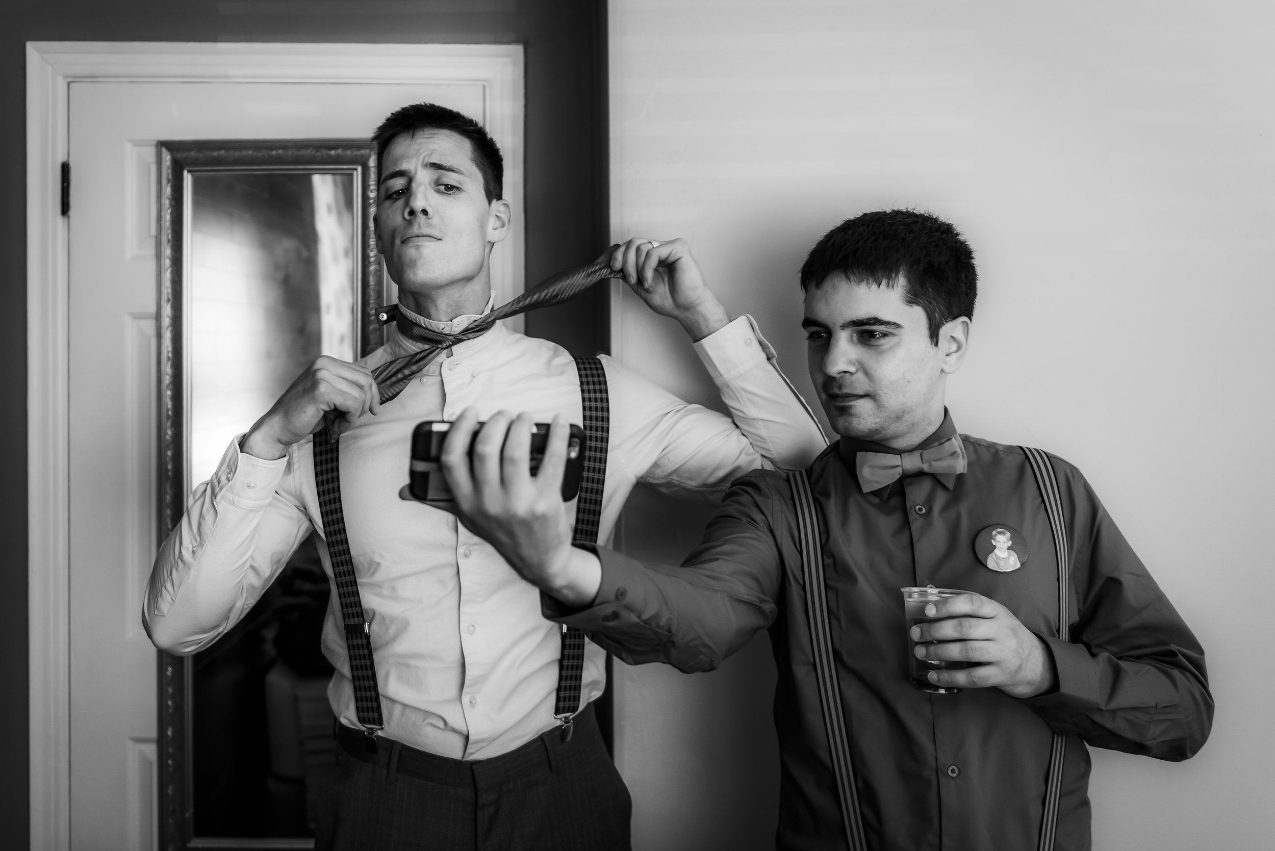 bestman helps groom with bowtie by holding phone video bride about to have first look with groom