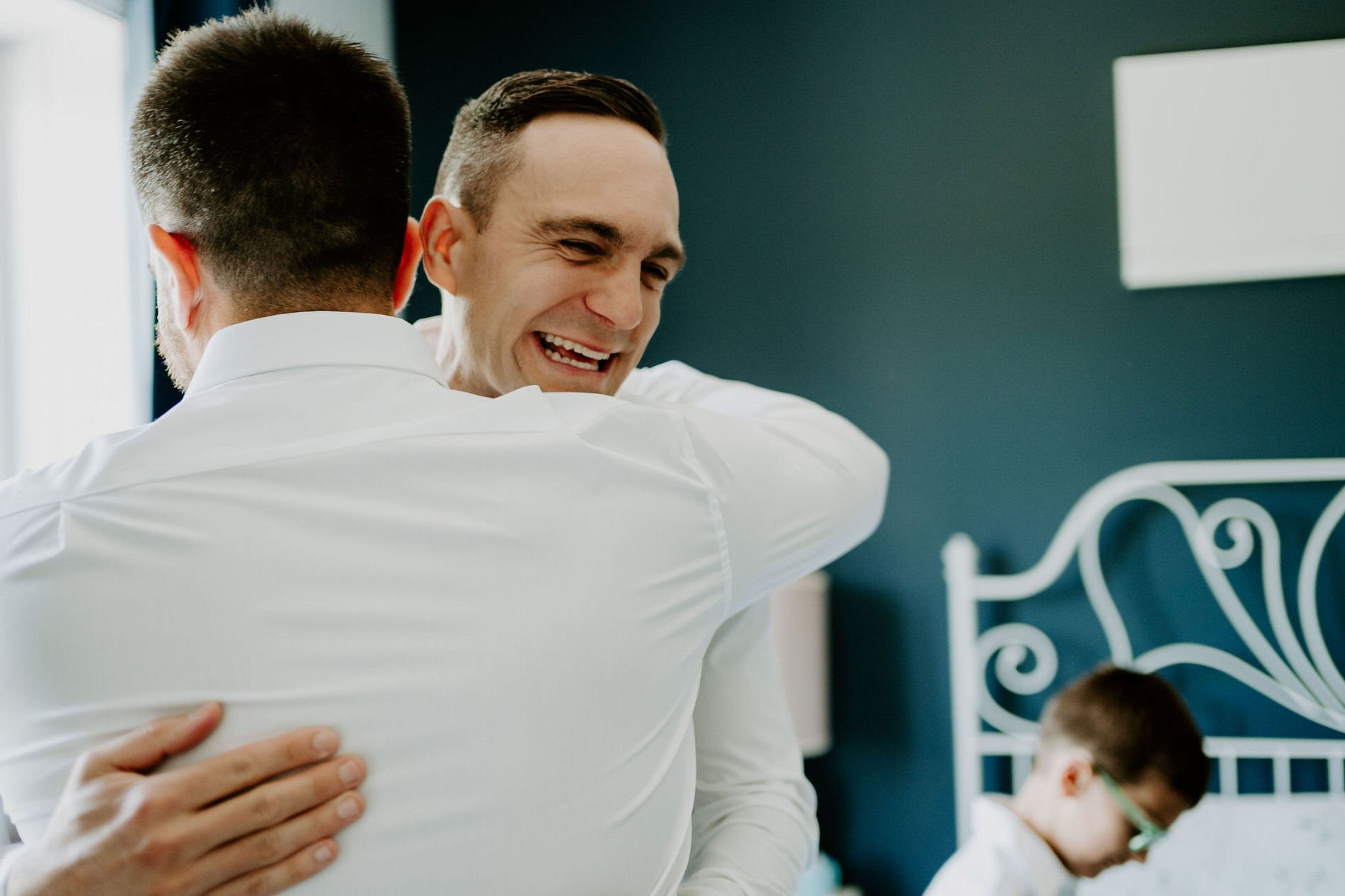 Petar beaming as he hugs a groomsman on the morning of his wedding, with a young boy on a bed in the background