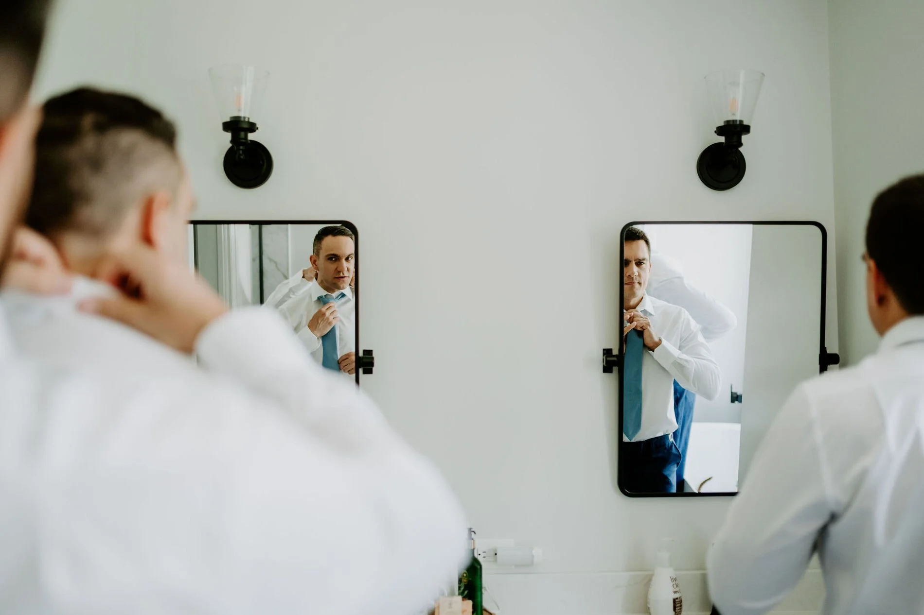Two men tying pale blue ties in twin bathroom mirrors before Holly and Petar's wedding