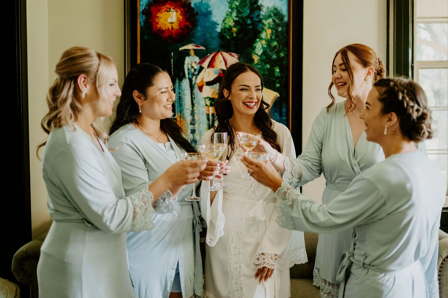 Holly in a white lace robe toasting champagne with four bridesmaids in sage green robes before her Evermore wedding