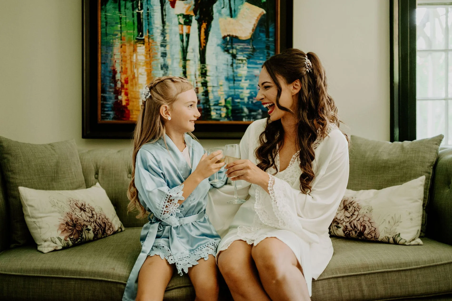 Holly and her flower girl laughing and clinking glasses on a green sofa as they get ready