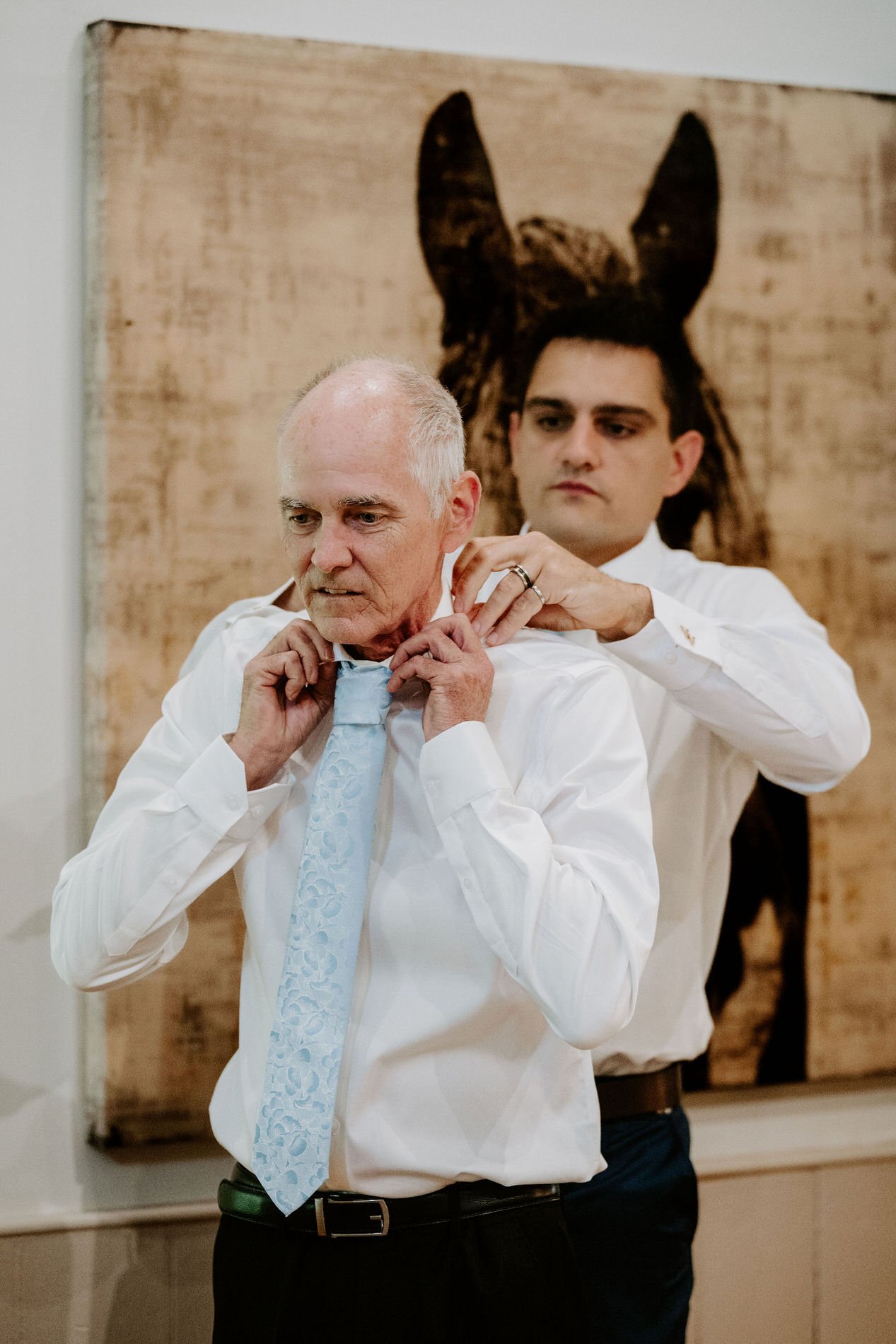 Young man fastening the collar of an older man's shirt and pale blue tie in front of a painting of a horse