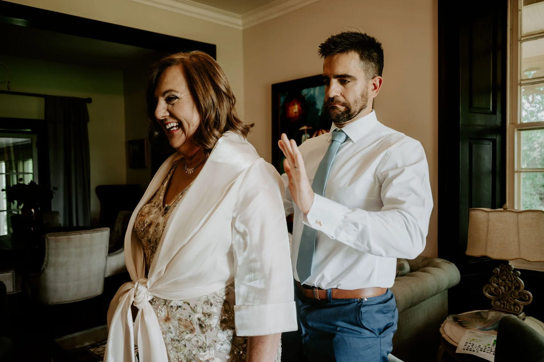 Man in a blue tie helping a laughing woman in a sequinned dress into a cream wrap on the wedding morning