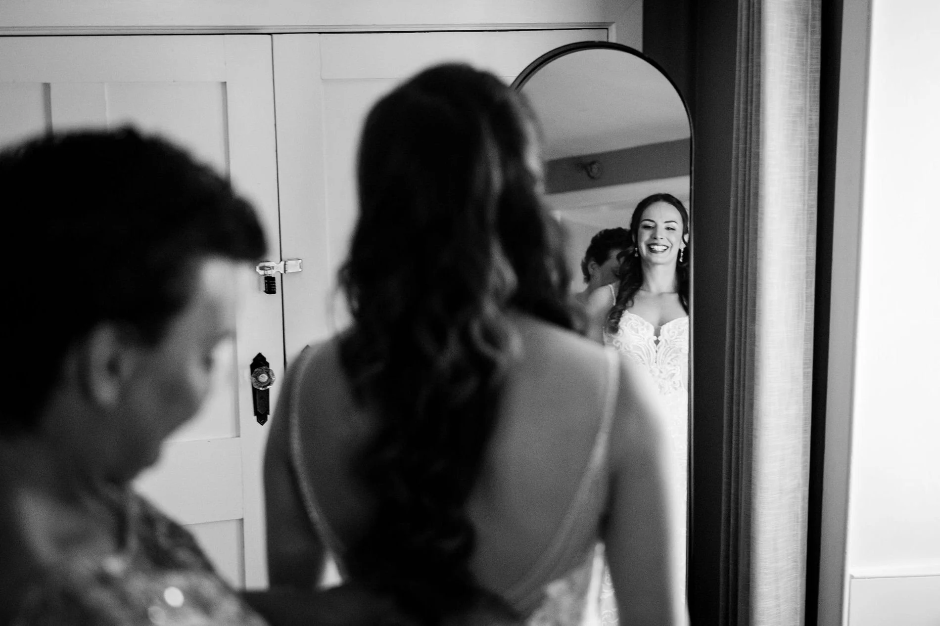 Black and white photo of Holly smiling at her reflection as a woman helps fasten her gown