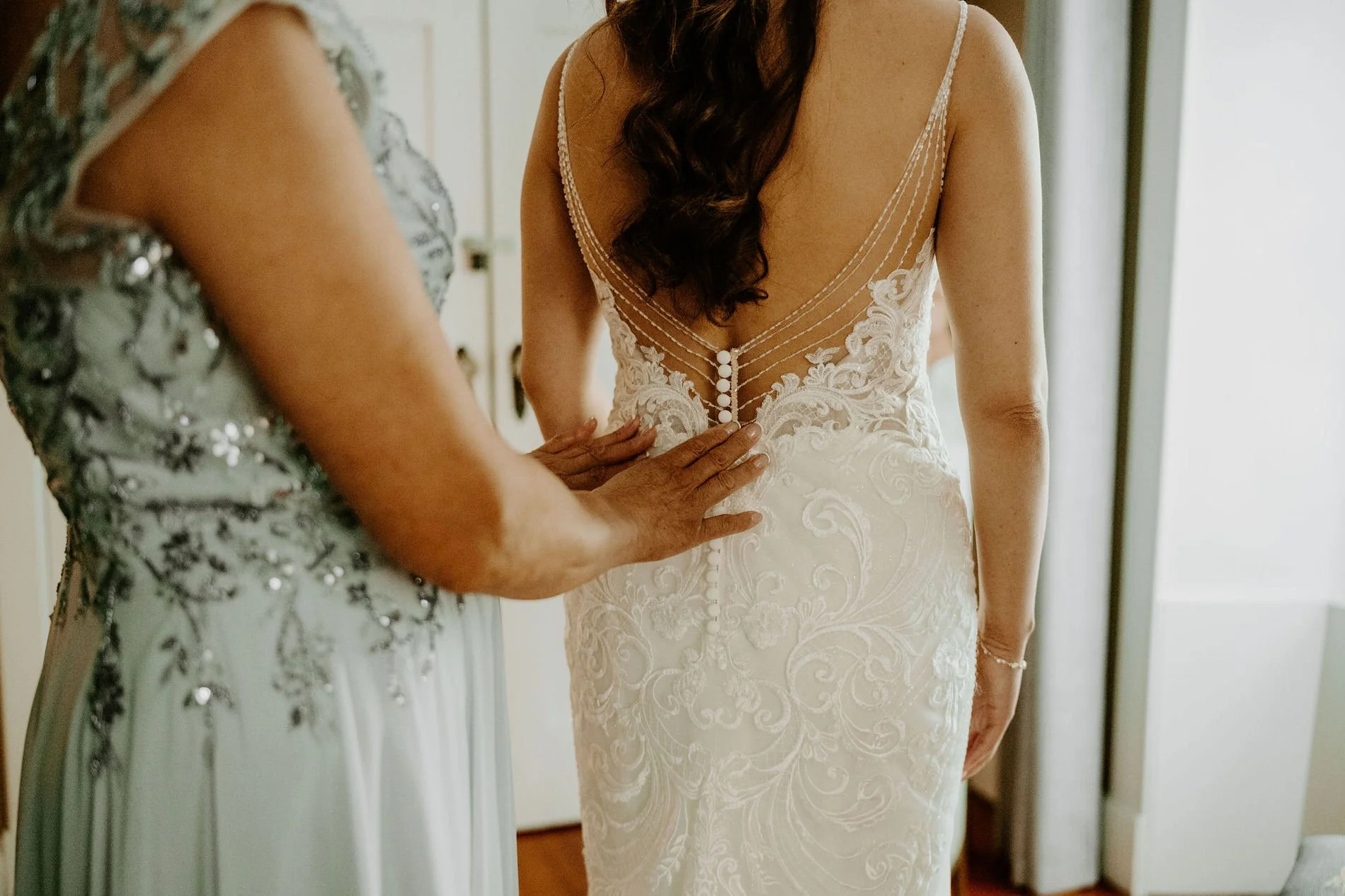 Woman in a sage sequinned dress buttoning the pearl buttons down the back of Holly's lace gown