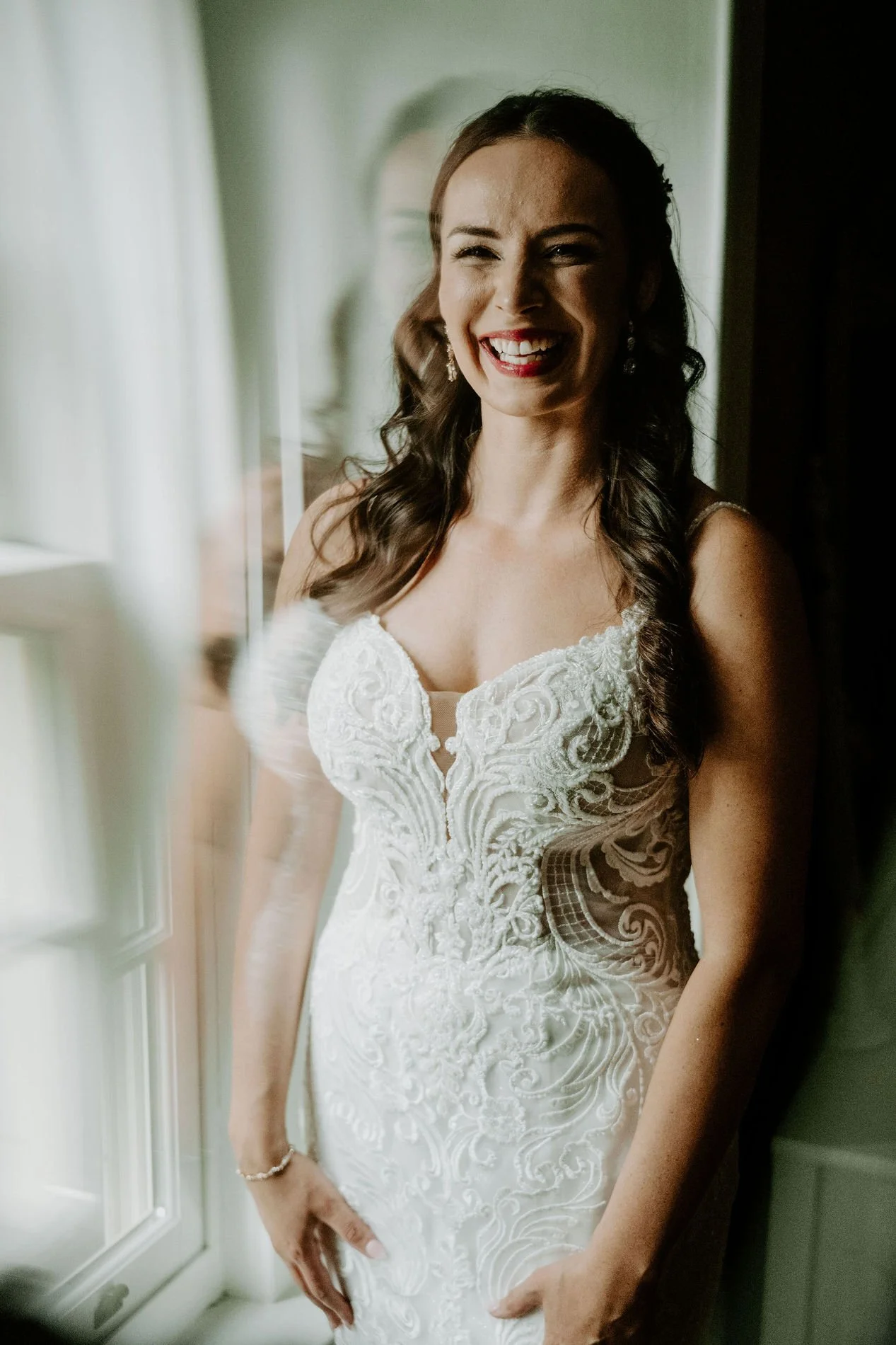 Holly laughing by a window in her fitted lace wedding gown