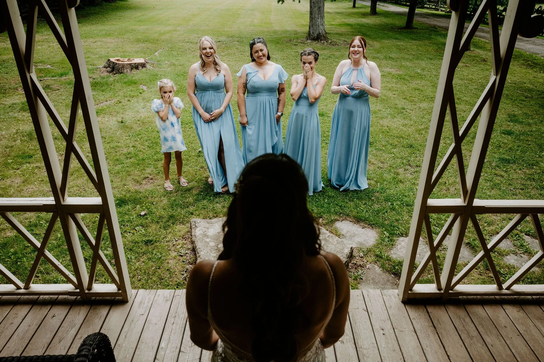 Holly on the porch at Evermore as her bridesmaids in dusty blue and the flower girl react with delight on the lawn