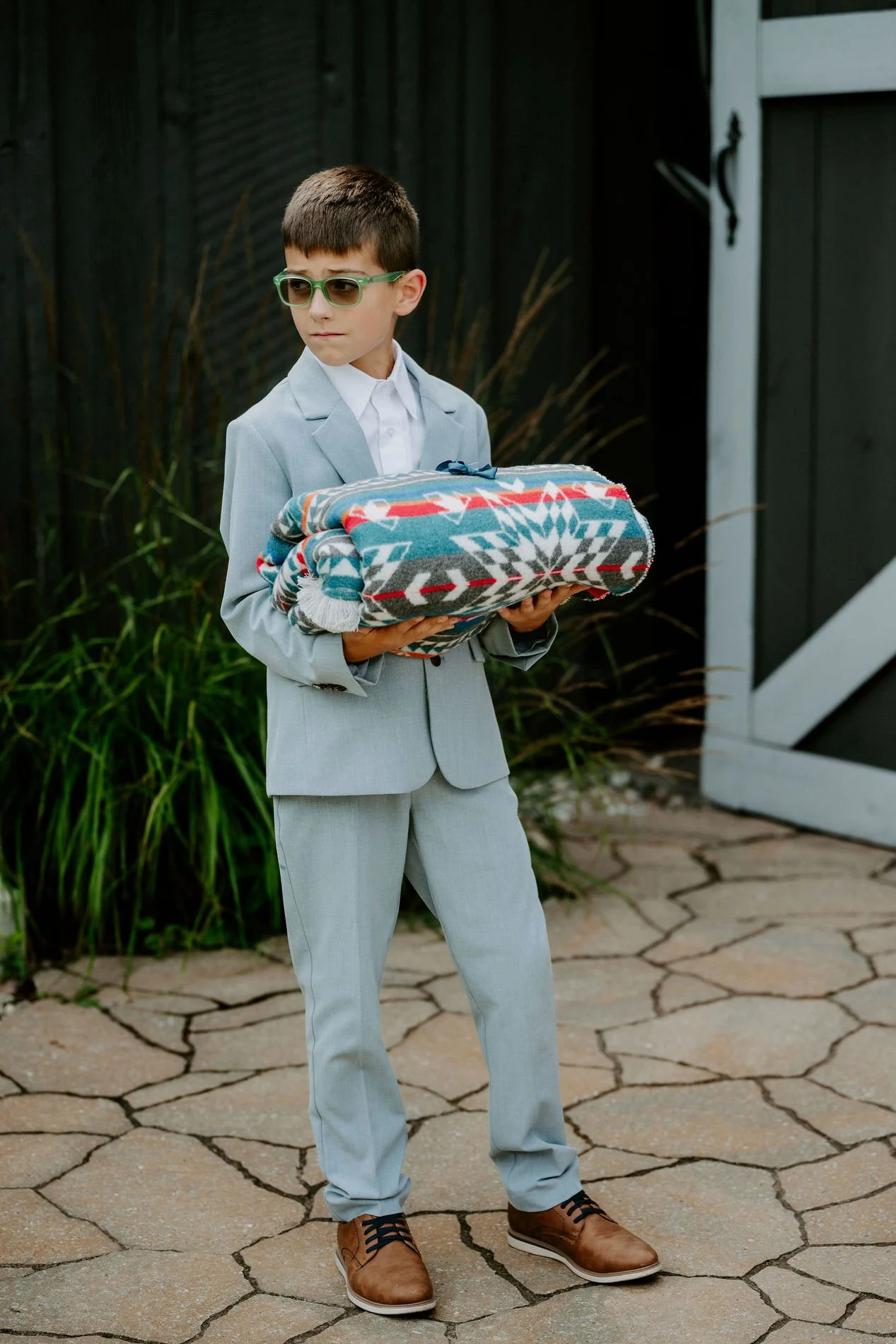 Young boy in a pale blue suit and green sunglasses carrying a rolled patterned blanket at Evermore
