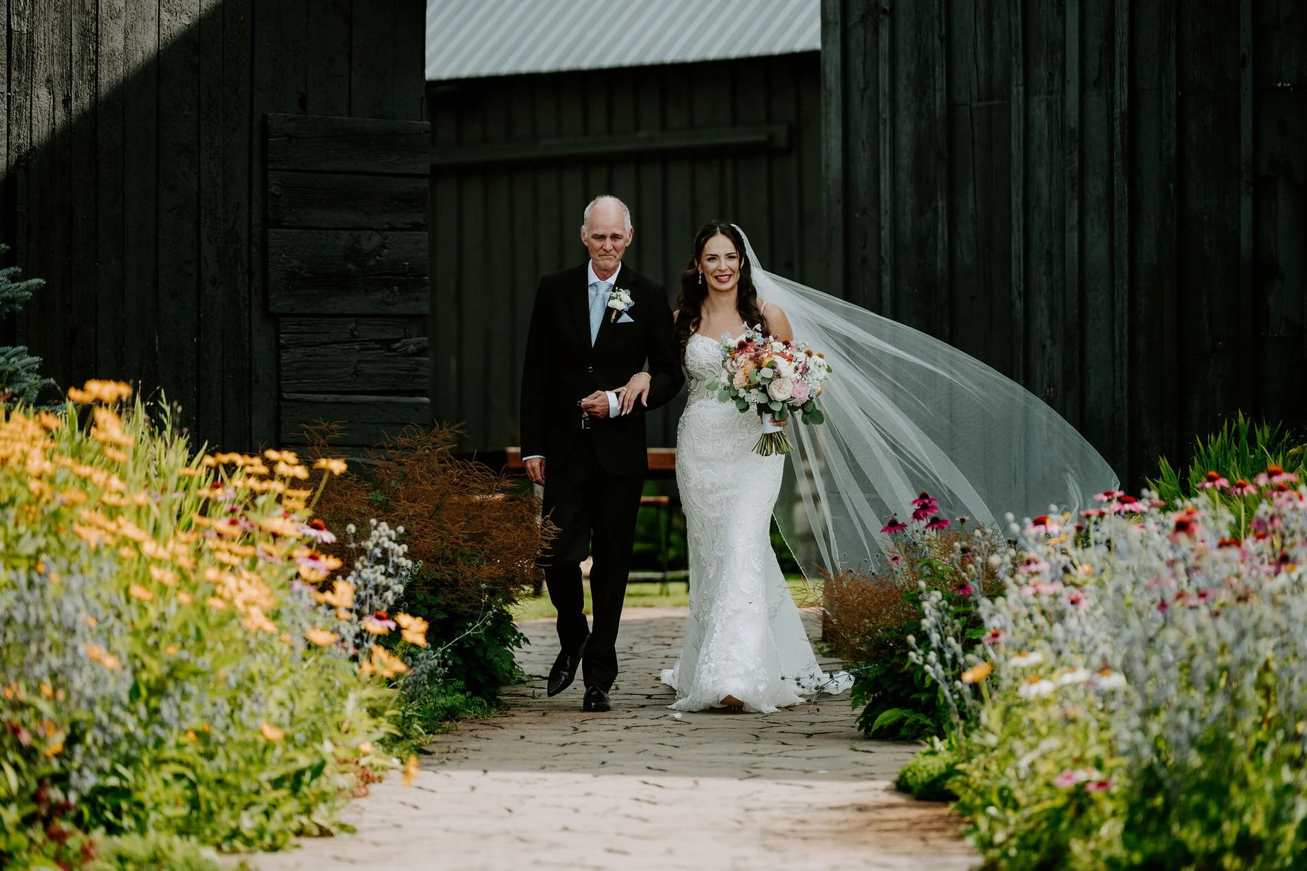 Holly's father walks her down the wildflower-lined garden path at Evermore, her long veil blowing behind her