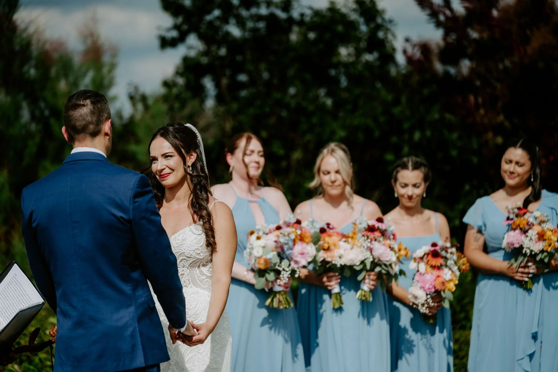 Holly smiling as she holds Petar's hands, bridesmaids in blue with bright bouquets behind her