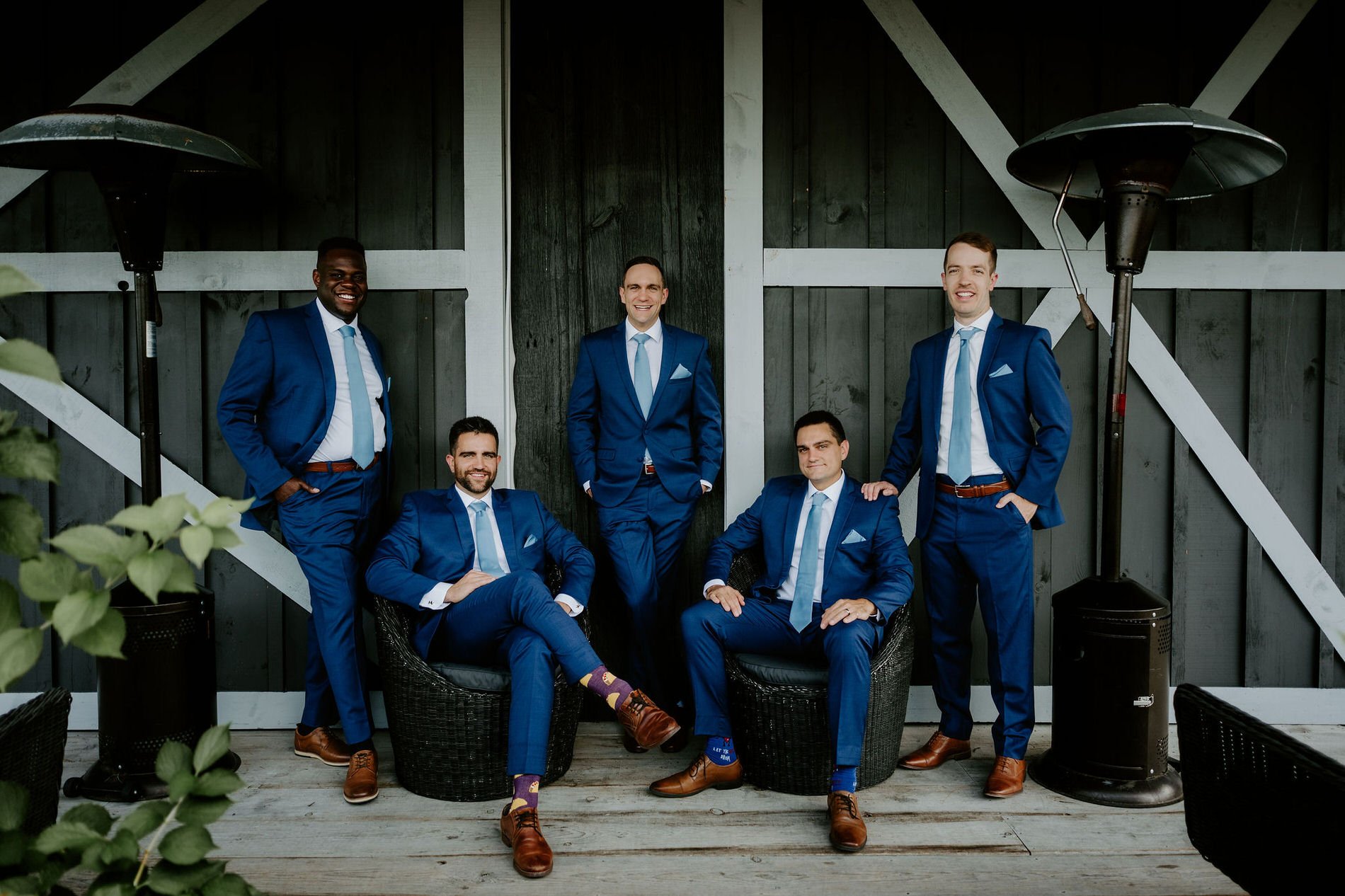 Groom and groomsmen in matching blue suits and colourful socks posing in front of a dark barn at Evermore