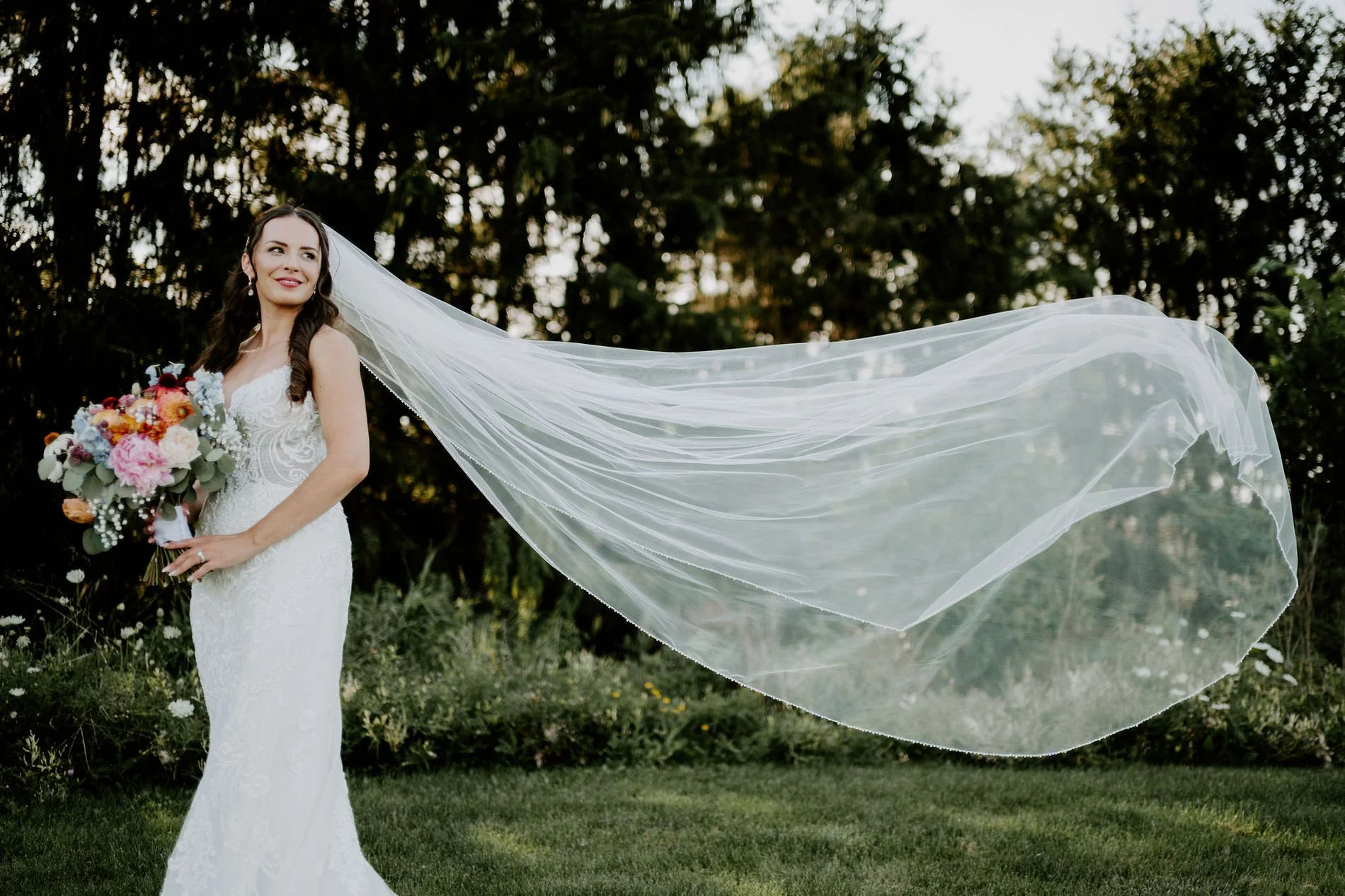 Holly holding her bouquet as her long cathedral veil billows out beside her in the gardens at Evermore