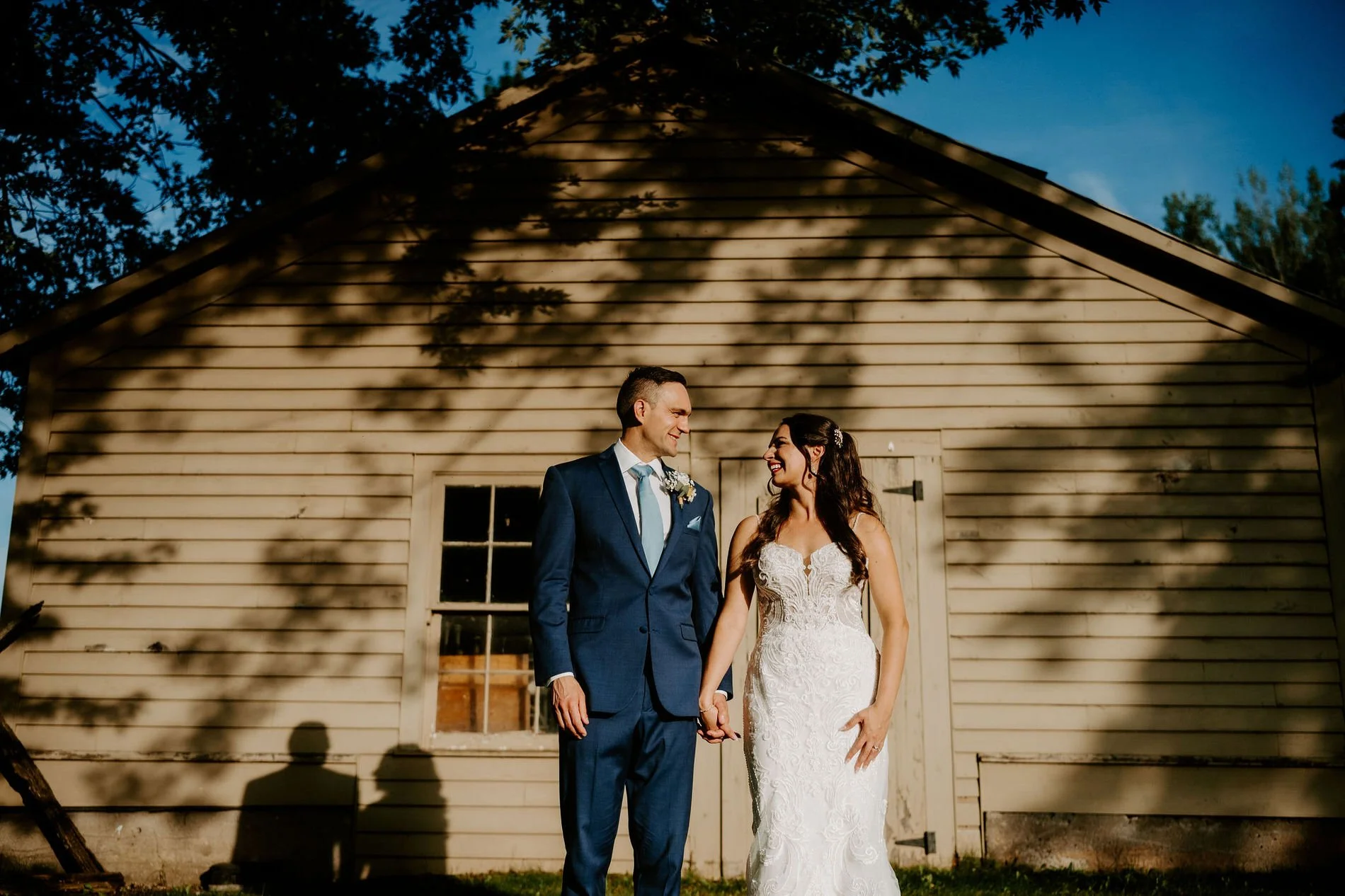 Holly and Petar holding hands and smiling at each other in front of a sunlit clapboard building at Evermore