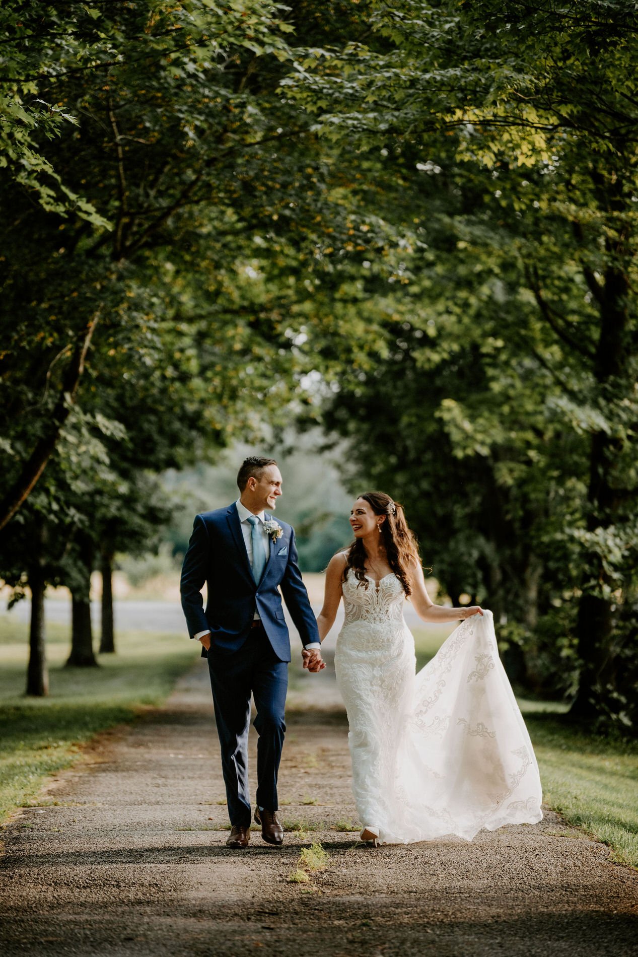Holly and Petar walking hand in hand along Evermore's tree-lined lane, Holly lifting her train