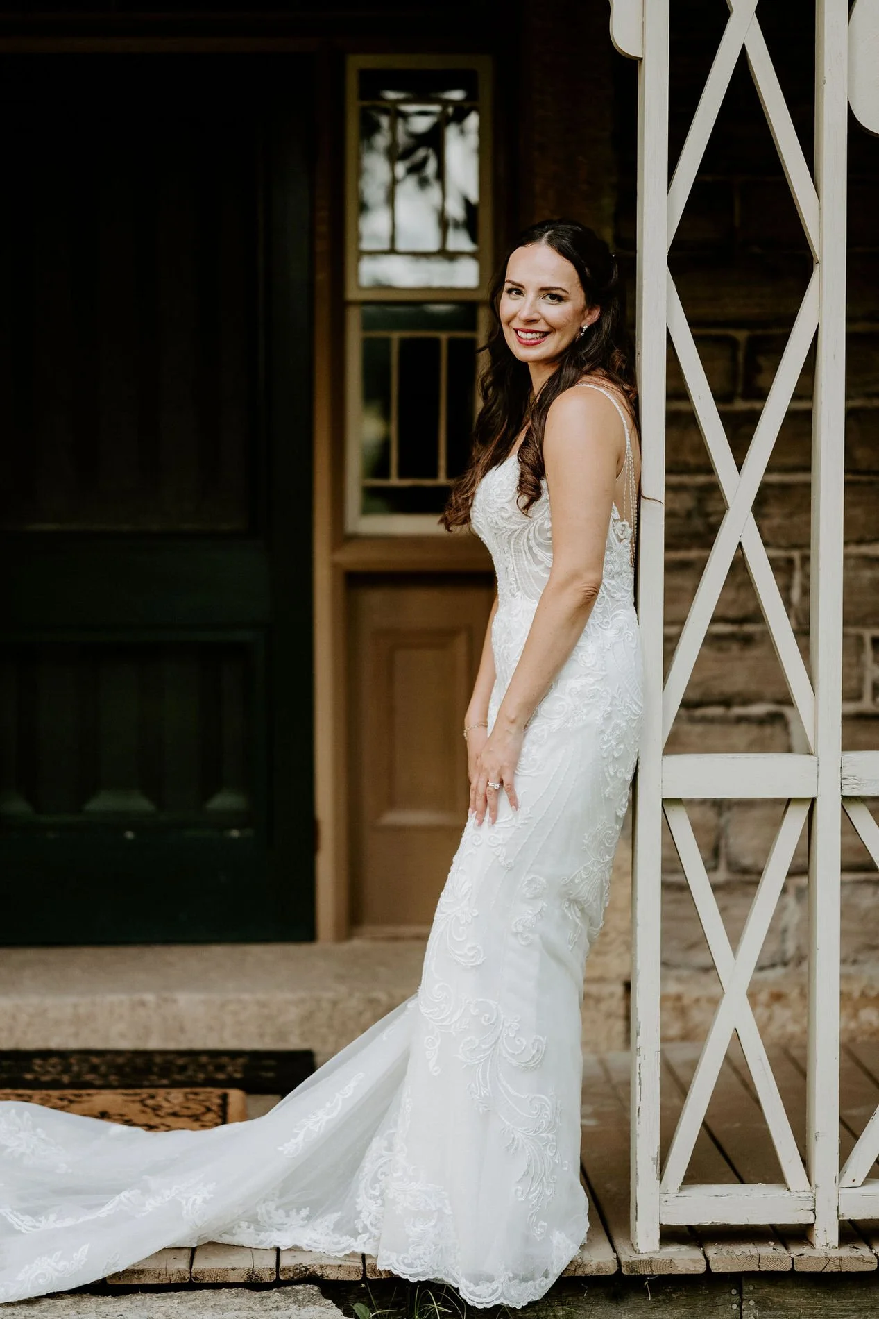 Holly smiling against a white lattice porch post at Evermore, her lace train spread across the boards