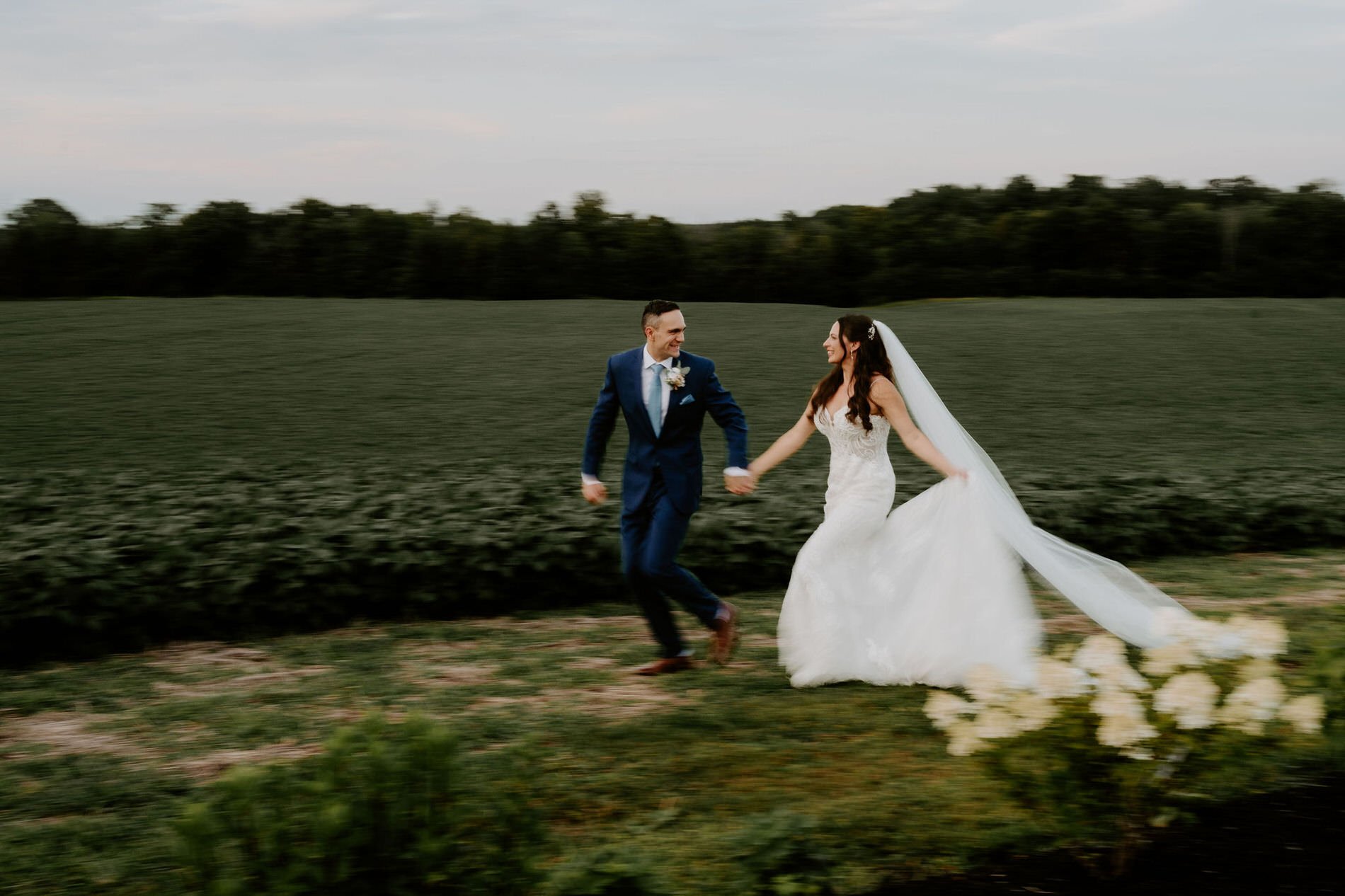 Holly and Petar running hand in hand along the edge of a field at Evermore, her veil trailing behind