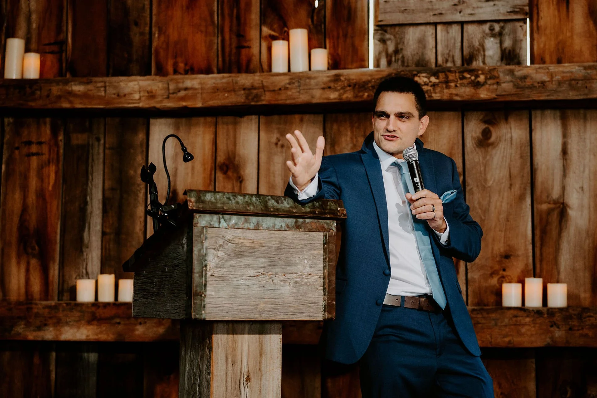 Man in a blue suit giving a speech at a rustic wooden lectern surrounded by candles in the barn at Evermore