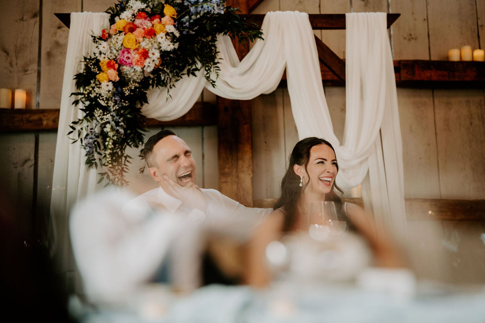 Holly and Petar laughing at the head table beneath draped fabric and a colourful floral arrangement