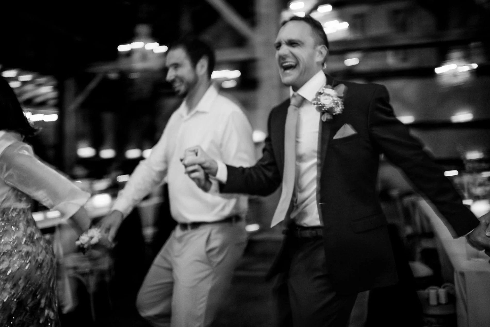 Black and white photo of a laughing man with a boutonnière leading a line of dancers