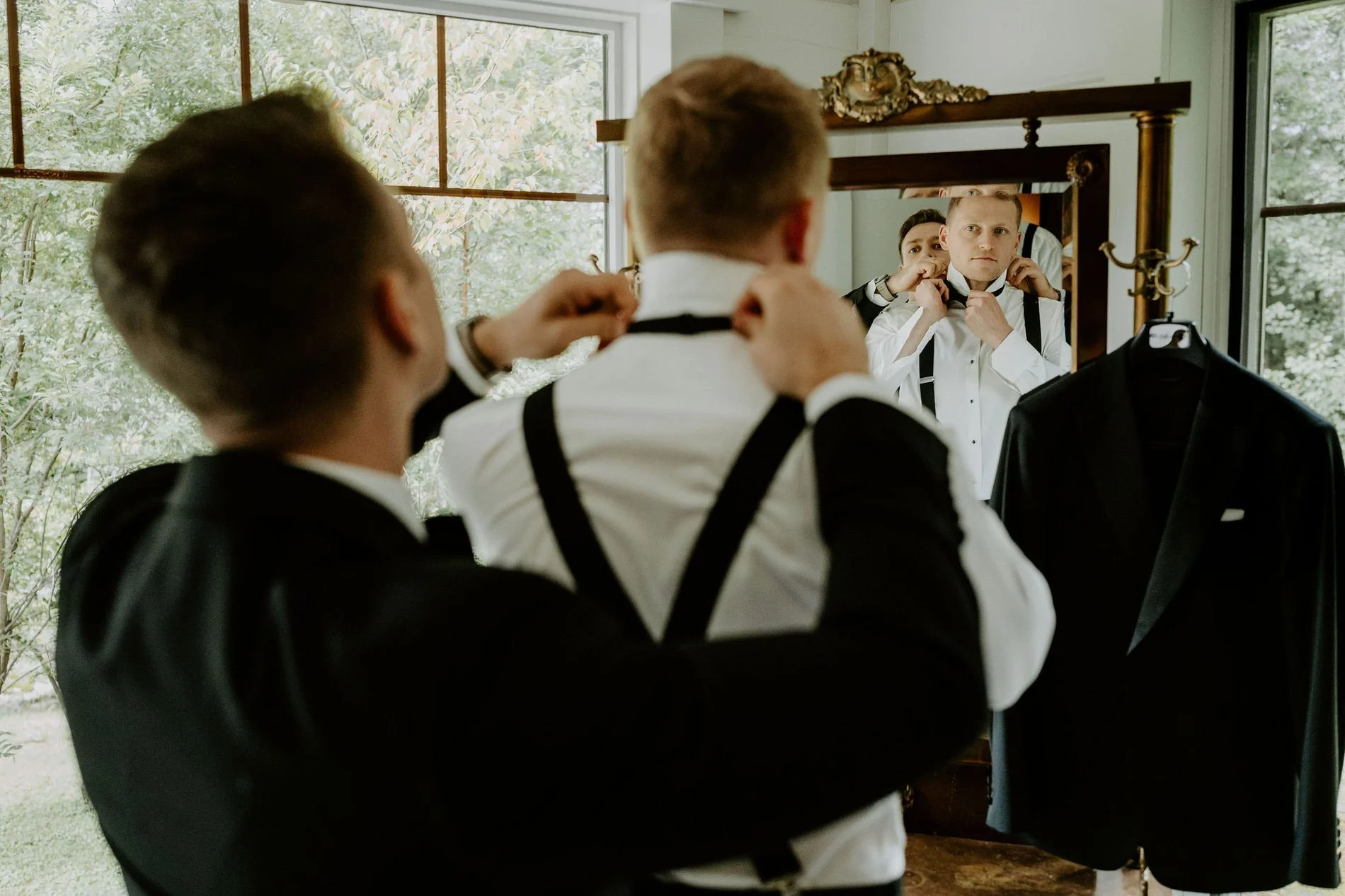 A groomsman fastening Will's bow tie as he watches in an ornate gilded mirror
