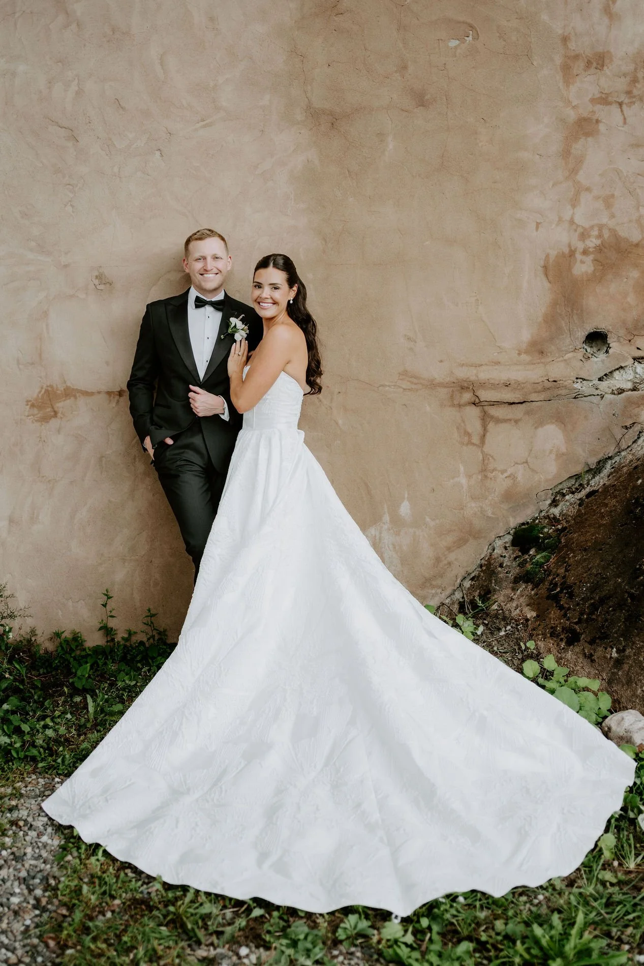 Kylie and Will against a textured plaster wall, her long train spread across the grass