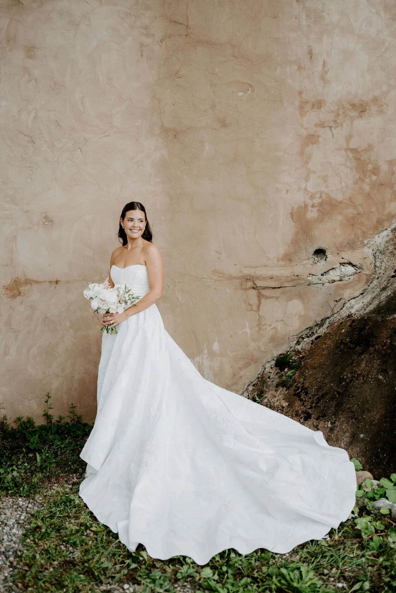 Kylie holding her bouquet, her dramatic train fanned out in front of a plaster wall at Le Belvedere