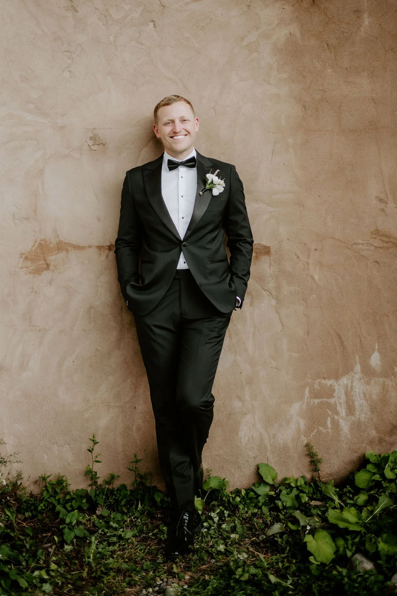Will leaning against a plaster wall in his black tuxedo, smiling with his hands in his pockets