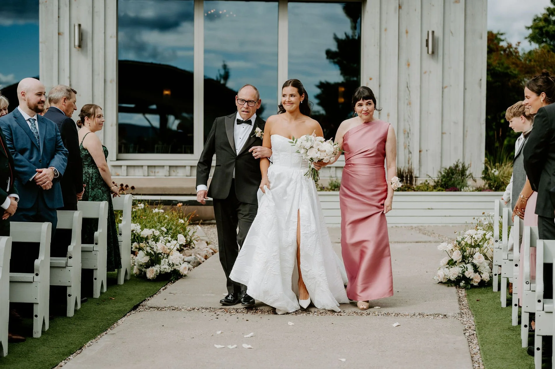 Kylie walking down the aisle at Le Belvedere between a man in a tuxedo and a woman in a rose-pink gown