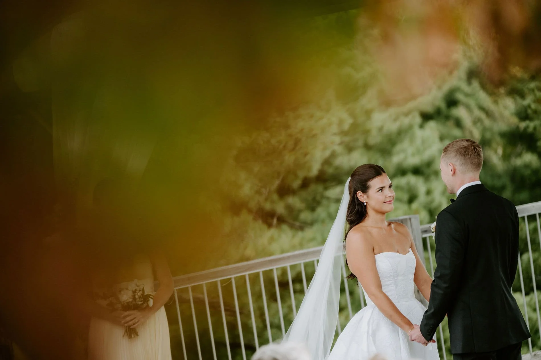 Kylie and Will holding hands during their ceremony, framed by blurred autumn leaves