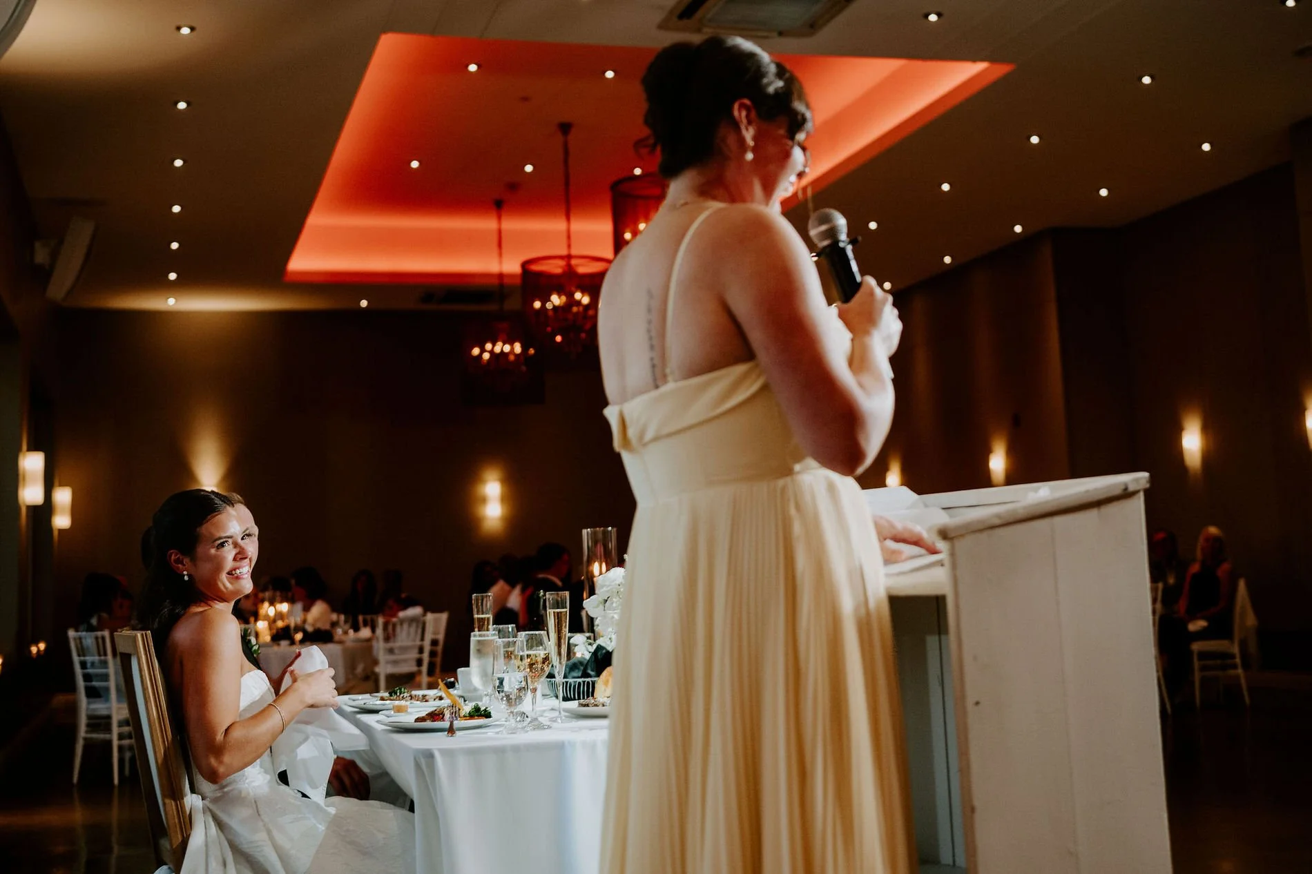 Kylie laughing at the head table as a bridesmaid in a pale yellow gown gives her speech
