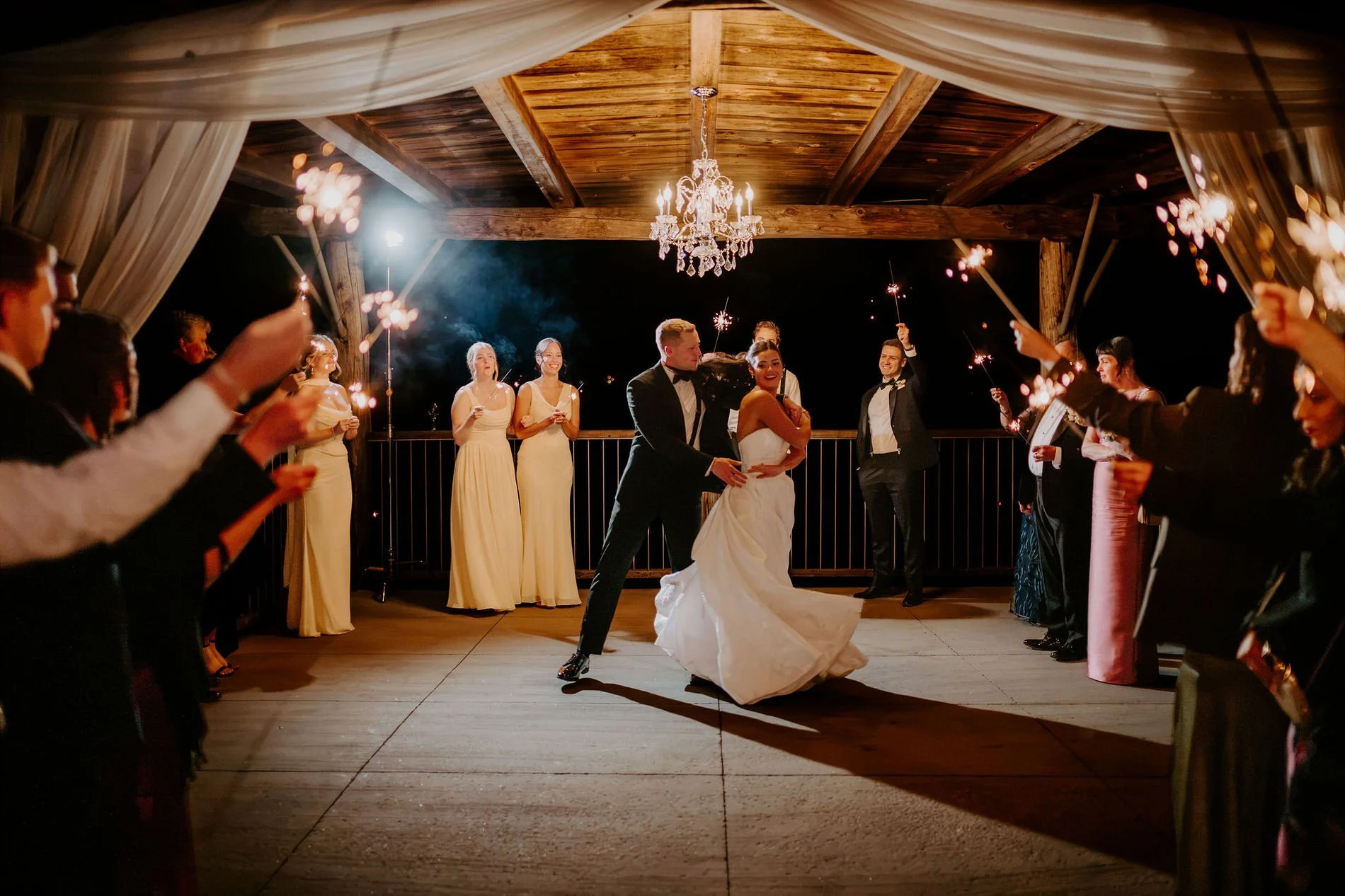 Kylie and Will dancing under the chandelier pavilion at night as the wedding party waves sparklers