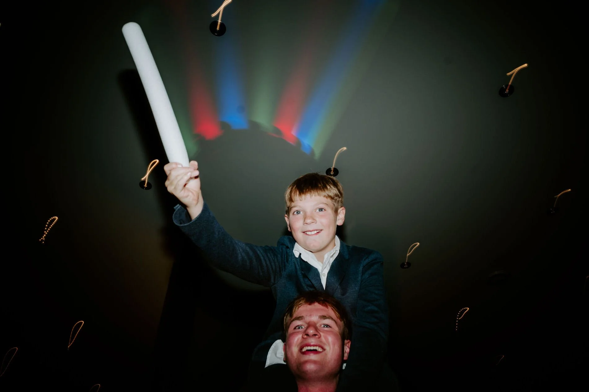A boy on a man's shoulders waving a foam glow stick under colourful dance-floor lights