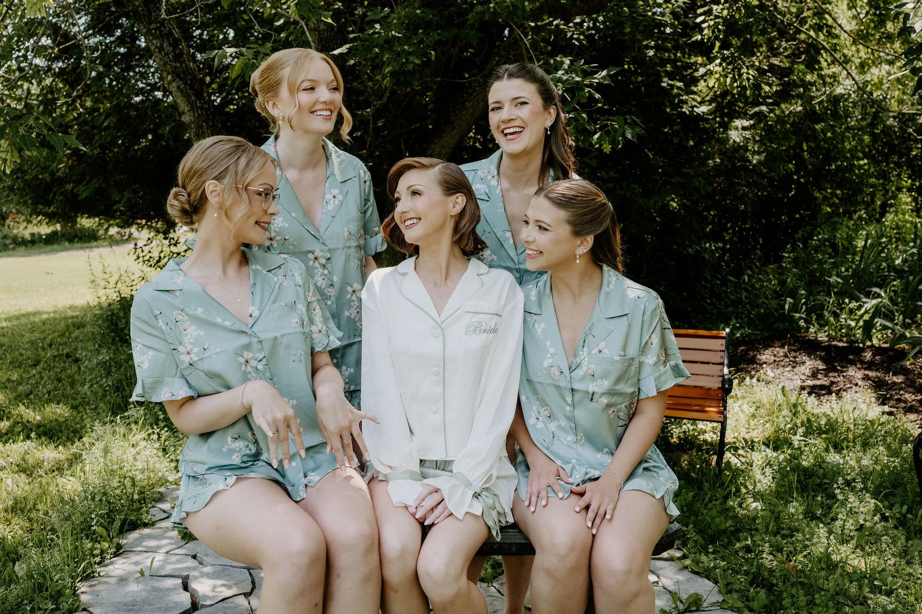 Emily laughing on a garden bench with four bridesmaids in matching sage floral pyjamas at Stonefields Estate