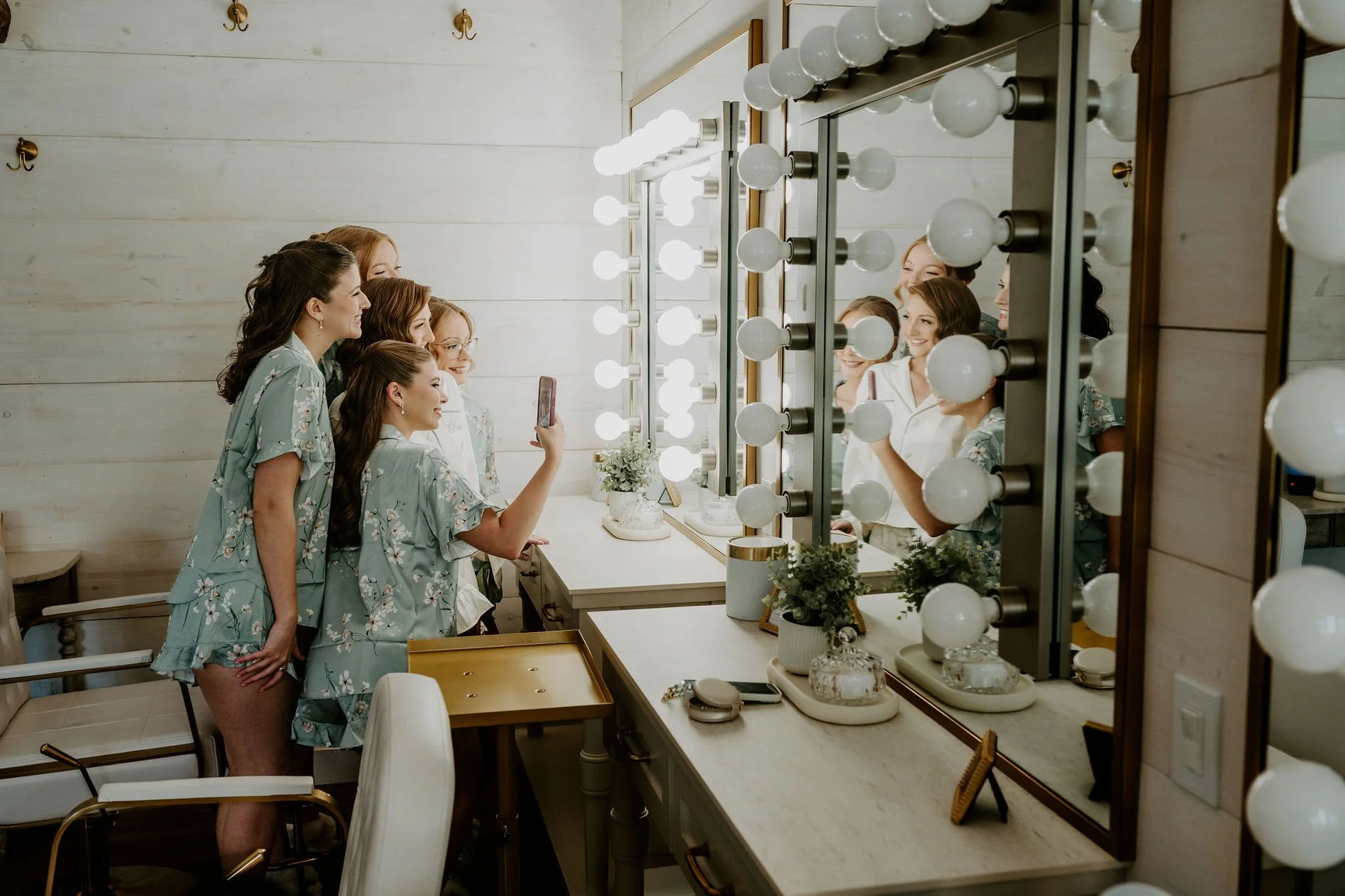 Bridesmaids in sage pyjamas taking a selfie in the bulb-lit vanity mirror of the bridal suite