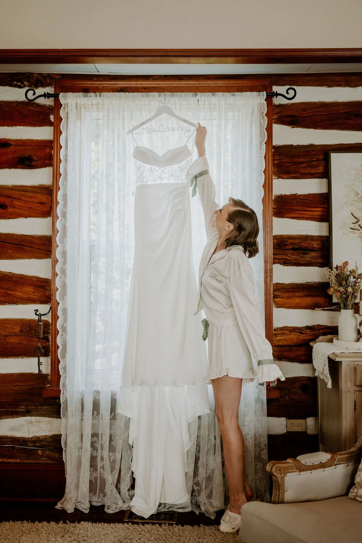 Emily hanging her wedding gown in a lace-curtained window of a log-walled room at Stonefields Estate