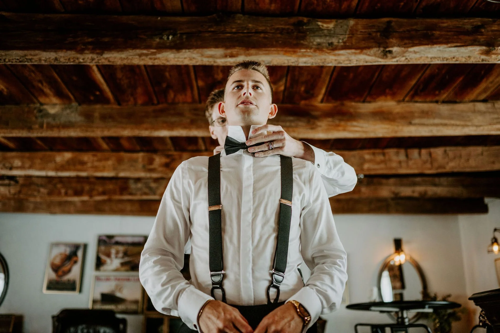 A groomsman fastening Austin's bow tie as he stands in white shirt and suspenders
