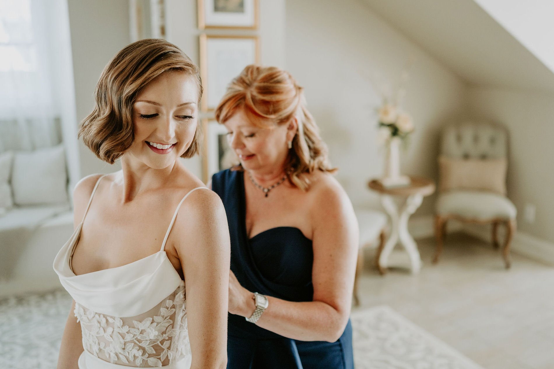 Emily smiling with her eyes closed as a woman in a navy gown fastens her dress