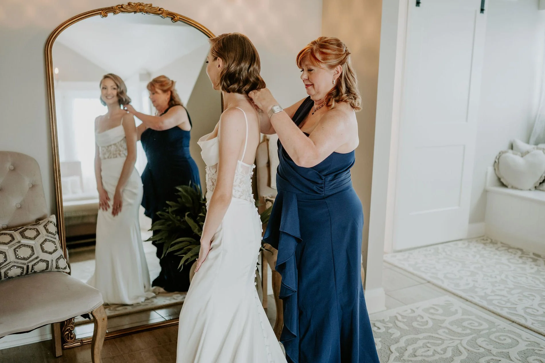 A woman in a navy gown fastening Emily's necklace in front of a tall gold-framed mirror