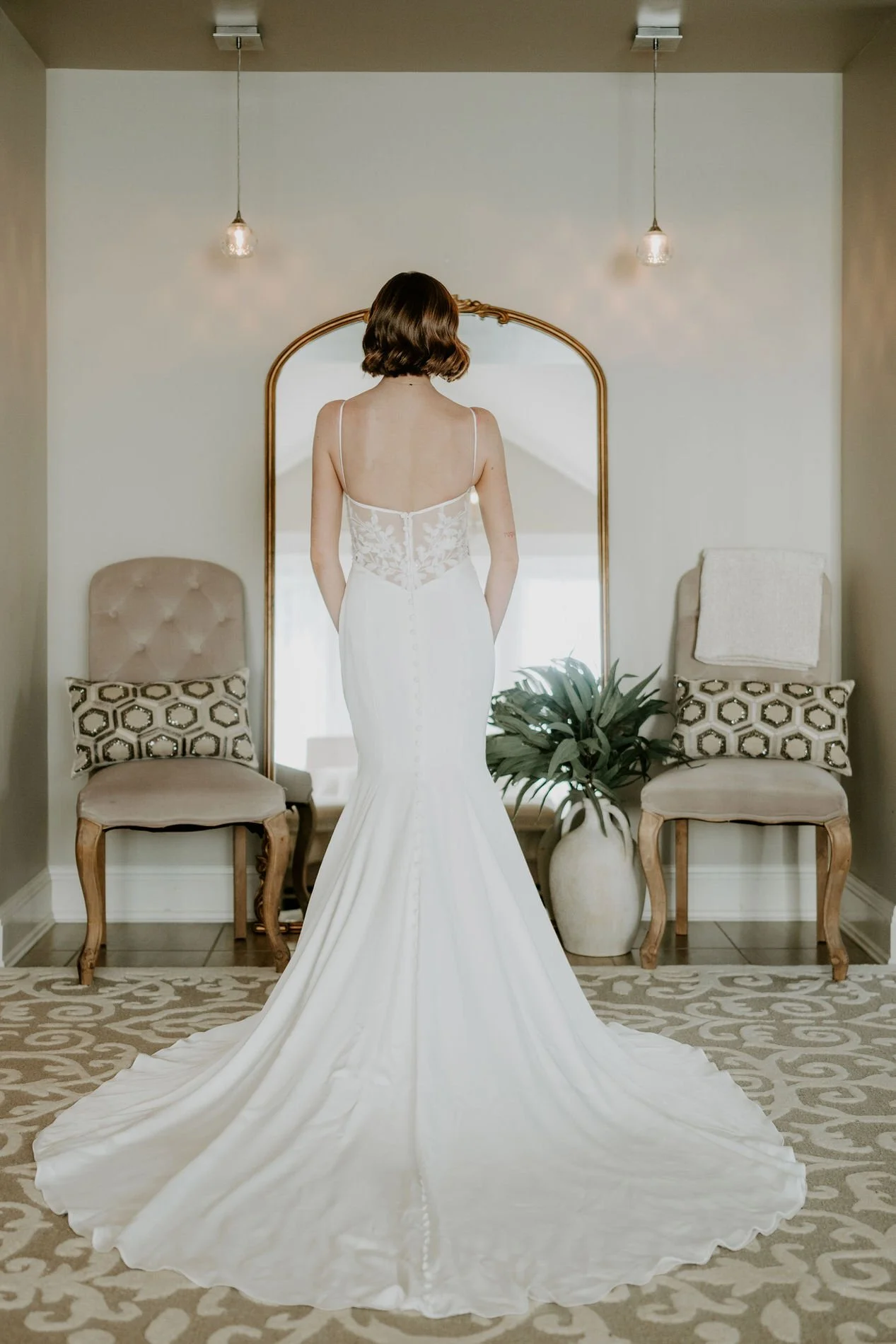 Emily from behind in her fitted gown with buttoned back and long train, facing a gold arched mirror