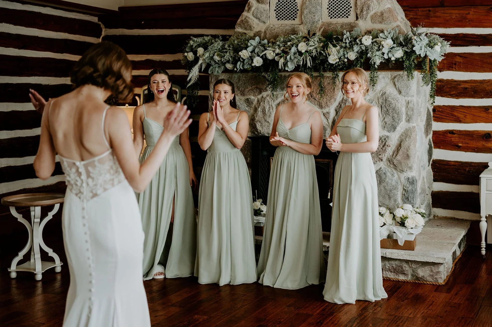 Bridesmaids in sage green gowns reacting with delight to Emily's dress beside a stone fireplace at Stonefields Estate